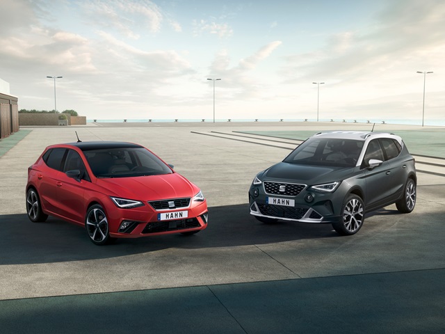Ein roter SEAT Ibiza steht neben einem grauen SEAT Arona. Beide Fahrzeuge sind in Frontansicht auf einem Parkplatz zu sehen. 
