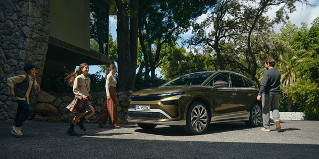 Škoda Enyaq Olibo-Grün Metallic in Front-Seitenansicht vor einem Haus mit mehreren Personen, darunter zwei Kinder und zwei Erwachsene, die auf das Fahrzeug zugehen