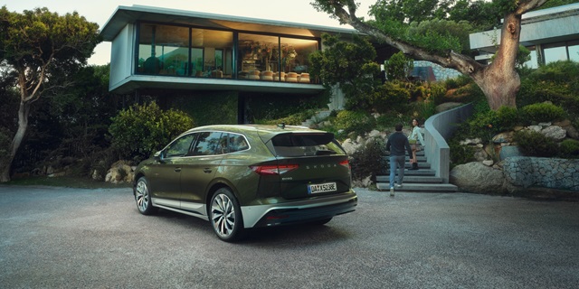 Škoda Enyaq Olibo-Grün Metallic in Heck-Seitenansicht vor modernem Haus mit großer Glasfront und zwei Personen auf einer Treppe