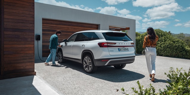 Weißer Skoda Karoq iV steht an einer Wallbox vor einem modernen Haus, zwei Personen nähern sich dem Fahrzeug
