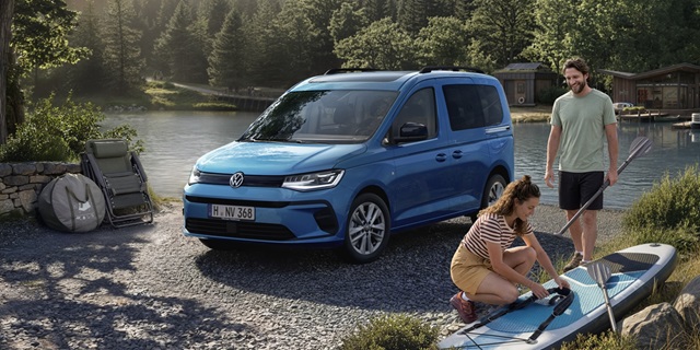 Blauer VW Caddy California steht am Ufer eines Sees, davor kniet eine Frau mit Paddelboard, ein Mann steht daneben mit Angelrute