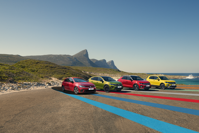 Vier bunte Volkswagen Modelle nebeneinander parkend auf einem großen Parkplatz an der Küste mit Bergkulisse im Hintergrund. 