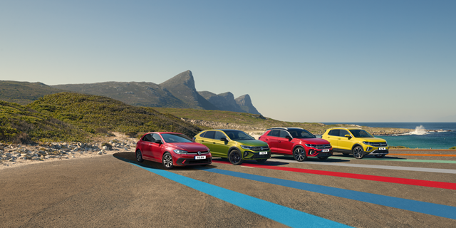 Vier bunte Volkswagen Modelle nebeneinander parkend auf einem großen Parkplatz an der Küste mit Bergkulisse im Hintergrund. 