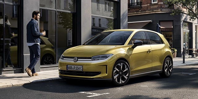 Seitenansicht eines gelben VW ID.Polo mit schwarzem Dach und markanten Felgen vor einem modernen Gebäude, daneben ein Mann mit Kaffeetasse im Gehen.