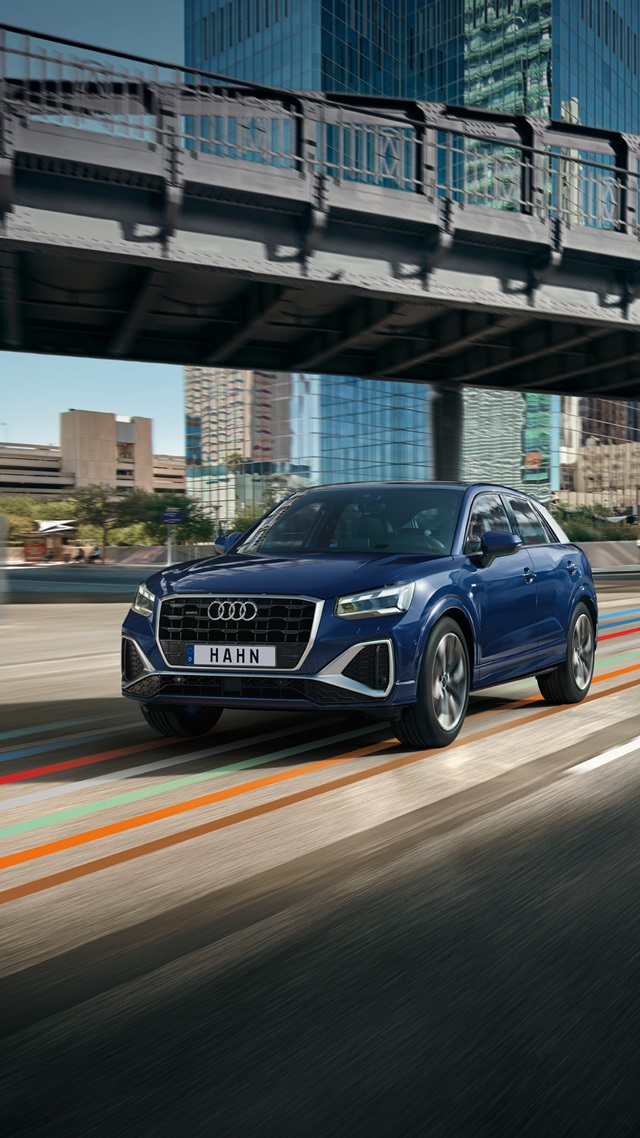 Blauer Audi Q2 fährt in Frontansicht auf einer Straße unter einer Brücke in urbaner Umgebung