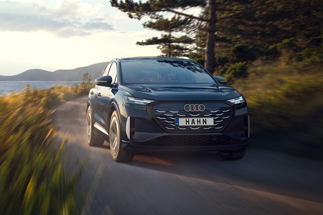Audi Q4 e-tron Sportback fährt auf einer Landstraße mit Bäumen und Bergen im Hintergrund, Frontansicht mit schwarzem Kühlergrill