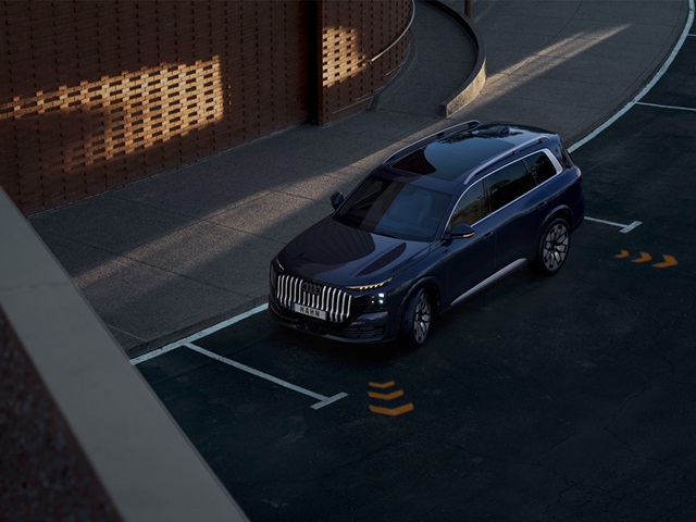 Blauer Audi Q9 steht in einer Parklücke vor einer Backsteinmauer mit sichtbaren Assistenzsystem-Sensoren an der Fahrzeugfront
