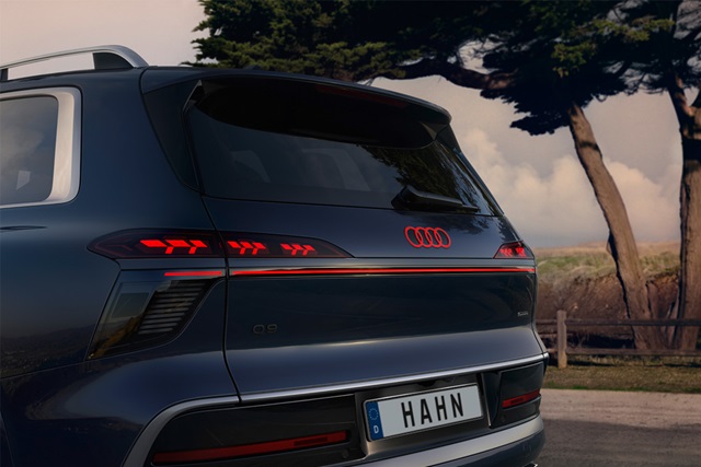 Heckansicht eines blauen Audi Q9 mit leuchtenden Rücklichtern und deutschem Kennzeichen 'HAHN' vor einer Landschaft mit Bäumen und Zaun