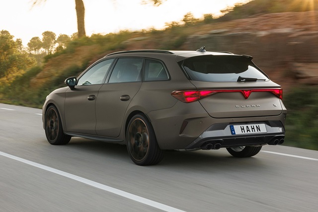 Heckansicht eines braunen CUPRA Leon Sportstourer mit durchgehendem Rücklicht und sportlichem Diffusor auf einer Straße mit Bäumen im Hintergrund