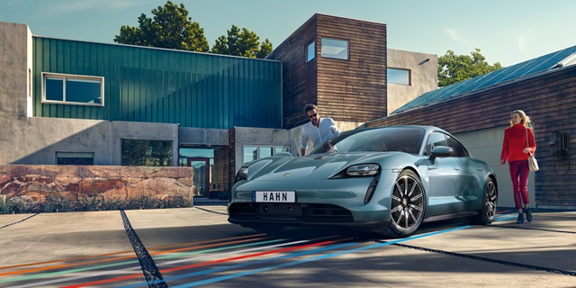 Der Porsche Taycan in blau vor einem Gebäude und in der Frontansicht. Auf der Fahrerseite steht eine Frau vor dem Auto und auf den Beifahrersitz nimmt gerade ein Mann Platz.