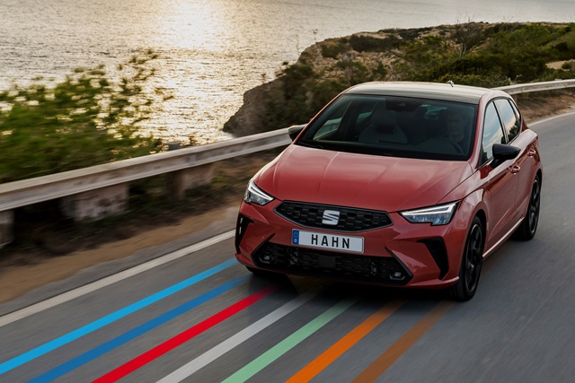 Roter SEAT Ibiza fährt auf Küstenstraße mit Wasser und Felsen im Hintergrund, Frontansicht mit schwarzem Kühlergrill und Nummernschild 'HAHN'