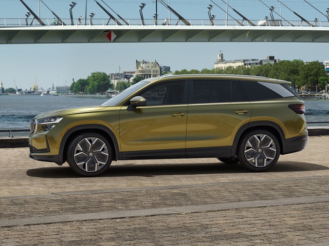 Seitenansicht eines gelben Škoda Kodiaq SUV vor einer Flussuferpromenade mit Brücke und Stadt im Hintergrund