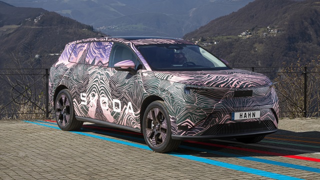 Škoda Peaq Erlkönig mit auffälliger Camouflage-Folie steht auf gepflastertem Parkplatz vor Berglandschaft.