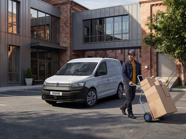Weißer VW Caddy Cargo vor einem modernen Backsteingebäude und ein Mann mit blauer Kappe und Jacke schiebt eine Sackkarre mit mehreren Kartons