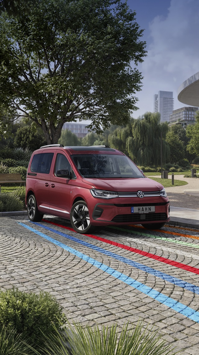 Roter VW Caddy steht auf gepflastertem Platz mit blauen und roten Linien, umgeben von Bäumen und moderner Stadtarchitektur im Hintergrund