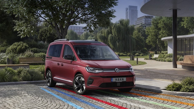 Roter VW Caddy steht auf gepflastertem Platz mit blauen und roten Linien, umgeben von Bäumen und moderner Stadtarchitektur im Hintergrund