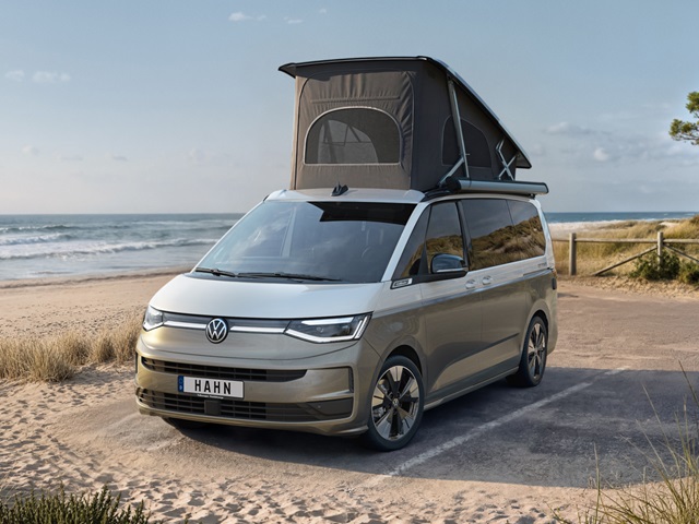 VW California Campervan in Beige-Weiß mit aufgeklapptem Dachzelt steht auf sandigem Strand mit blauem Himmel