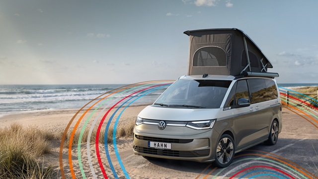VW California Campervan in Beige-Weiß mit aufgeklapptem Dachzelt steht auf sandigem Strand mit blauem Himmel