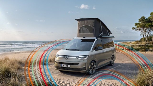 VW California Campervan in Beige-Weiß mit aufgeklapptem Dachzelt steht auf sandigem Strand mit blauem Himmel