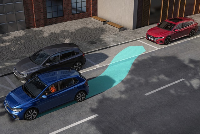 Blauer VW Polo parkt rückwärts in eine Parklücke mit sichtbarem blauen Assistenzpfad auf der Straße neben einem roten Auto und einem Gebäude mit Backsteinfassade