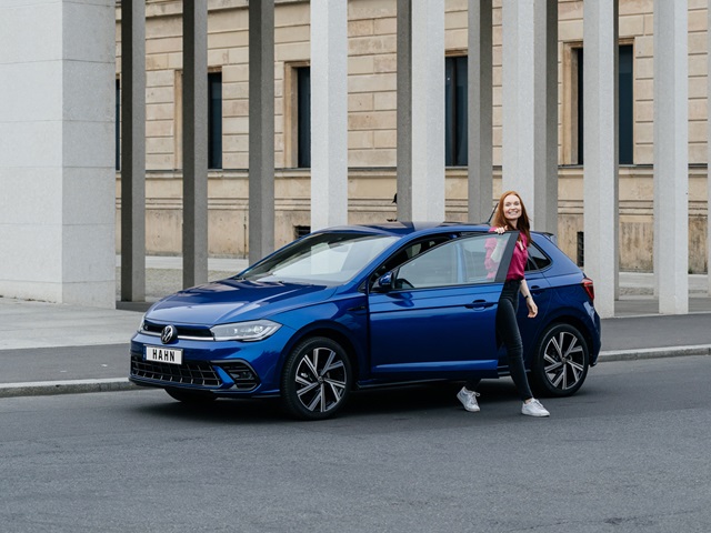 Blauer VW Polo steht seitlich vor einem Gebäude mit Säulen, eine Frau öffnet die Fahrertür und lächelt