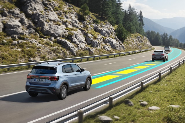 VW T-Cross fährt auf einer Landstraße mit farbigen Markierungen auf der Fahrbahn, weitere Fahrzeuge voraus, Berge und Bäume am Straßenrand.