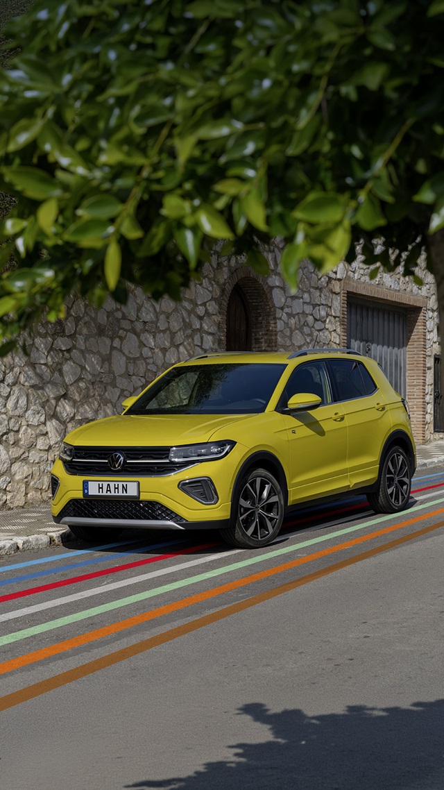 VW T-Cross R-Line in Gelb aus schräger Frontansicht auf einer Straße mit bunten Streifen, im Hintergrund Steinmauer und Tor.