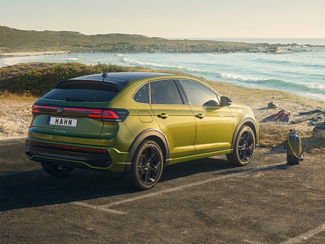 Grüner VW Taigo R-Line steht auf Asphaltstraße mit Meer und Küstenlandschaft im Hintergrund, daneben steht ein Rucksack