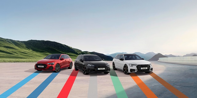 Audi Gebrauchtwagen :plus Wochen – Drei sportliche Audi-Fahrzeuge mit den bunten, dynamischen Linien des Hahn Codes auf einem Parkplatz vor einer Bergkulisse.