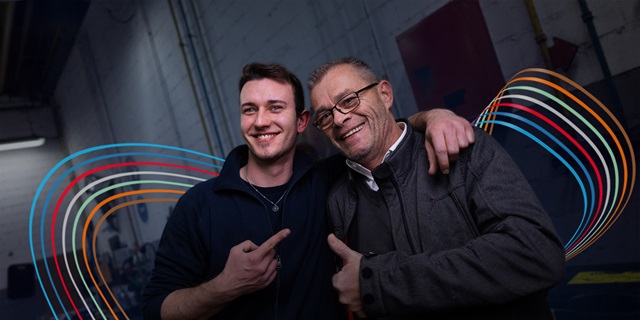 Zwei Männer posieren fröhlich für ein Teamfoto mit den bunten Linien des Hahn Codes im Hintergrund.