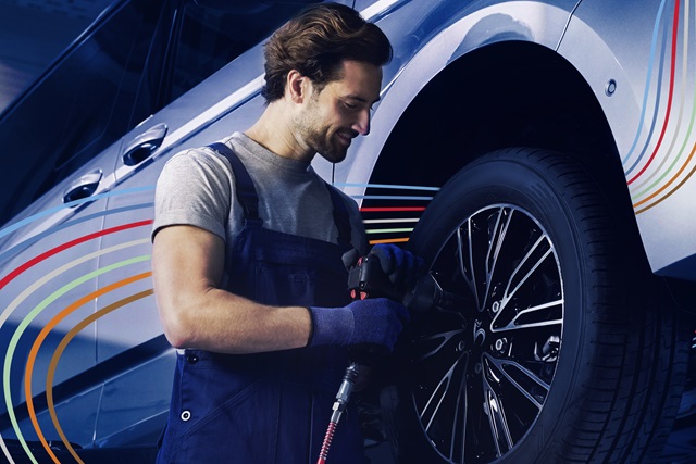 Mann in blauer Arbeitskleidung montiert mit einem Werkzeug einen Reifen an einem Auto.
