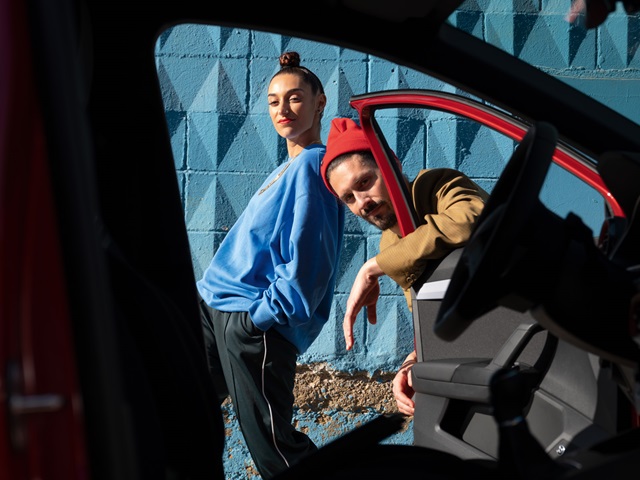 Ein Mann und eine Frau stehen vor einer blauen Wand und werden aus einem Auto heraus fotografiert