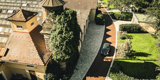 Luftaufnahme der Alten Weberei mit einem gepflasterten Weg, einem alten Gebäude mit Schornstein und einem geparkten Oldtimer auf dem Weg.