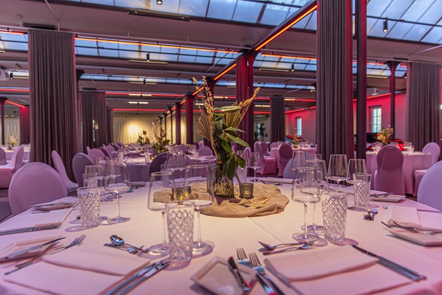 Festlich gedeckte runde Tische mit Gläsern, Besteck und Blumenarrangements in großen Saal der alten Weberei.