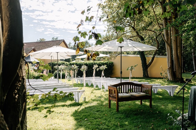 Garten der alten Weberei mit weißen Bänken und großen Sonnenschirmen, umgeben von Bäumen und grüner Wiese.