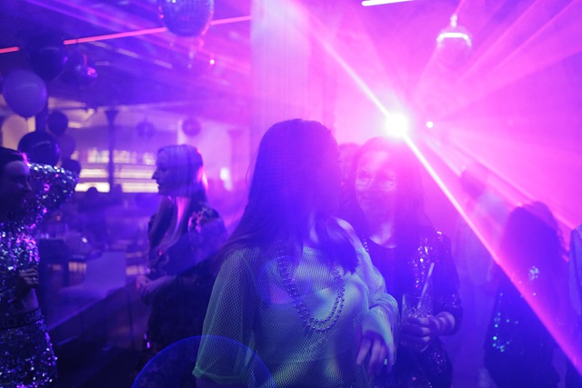 Menschen auf einer Party mit buntem Licht und Nebel, eine Person im Vordergrund trägt eine Kette.