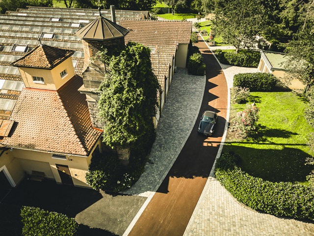 Luftaufnahme der Alten Weberei mit einem gepflasterten Weg, einem alten Gebäude mit Schornstein und einem geparkten Oldtimer auf dem Weg.