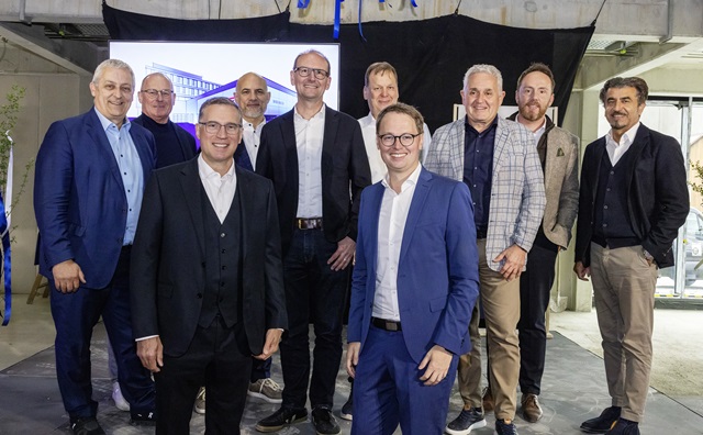 Gruppe von neun Männern in Anzügen und Sakko in einem unfertigen Innenraum mit Betonwänden und -boden
